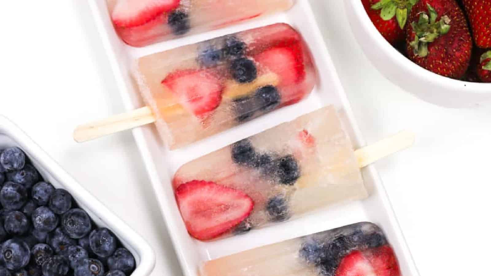 prosecco popsicles with strawberries and blueberries on a white ceramic tray next to bowls of fresh berries.
