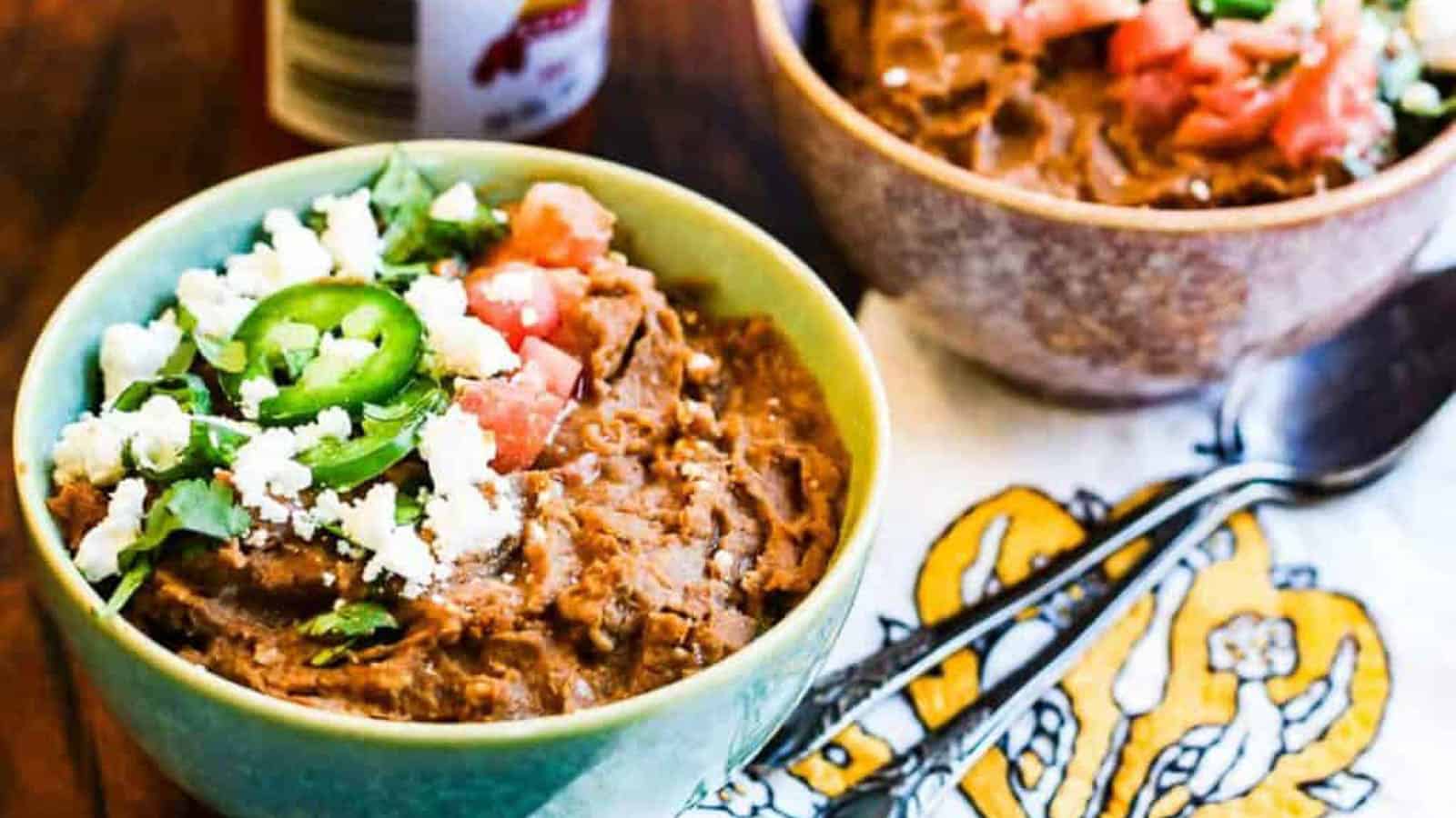 Bowls of refried beans garnished with sliced jalapenos, cheese, and tomatoes with a napkin and two spoons on the side.