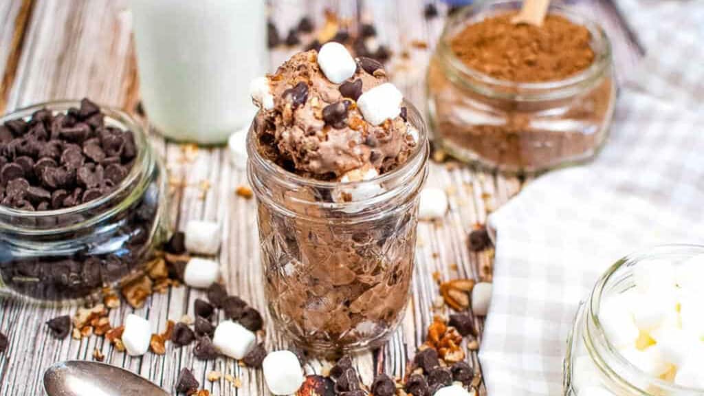 Low angle shot of rocky road ice cream in a canning jar with chocolate chips, marshmallows and nuts scattered about.