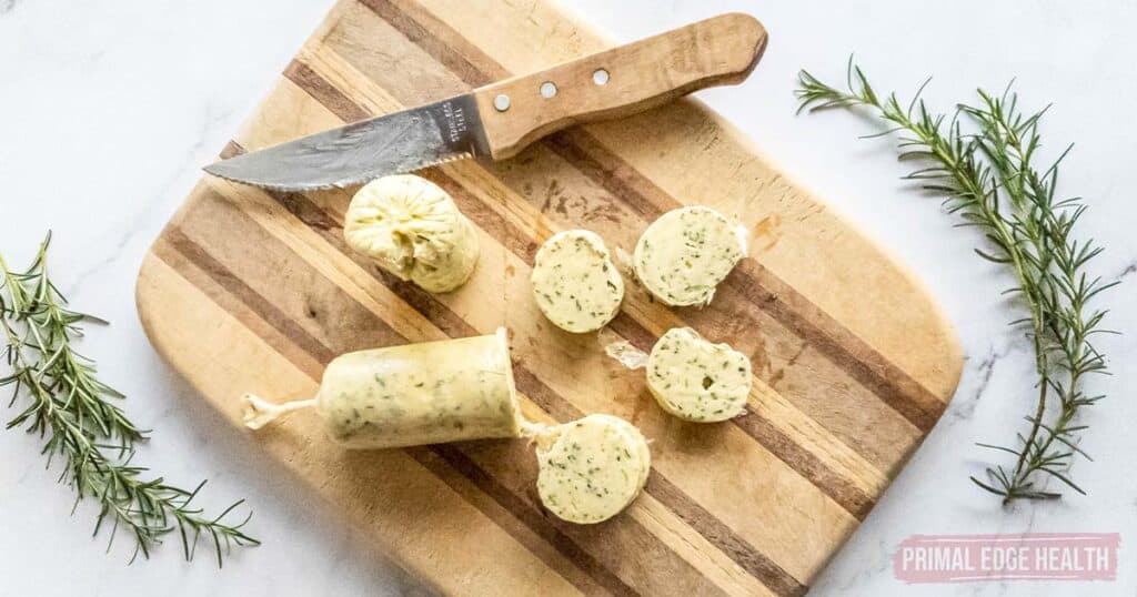 Rosemary garlic compound butter sliced on cutting board with kinfe and fresh herb.