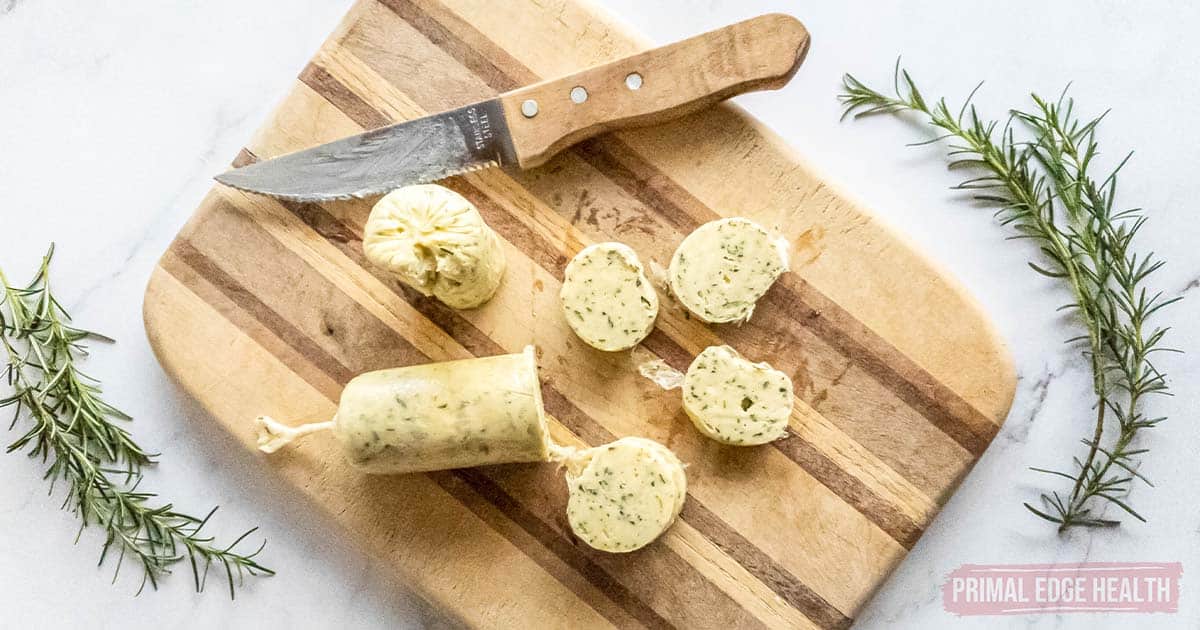 Rosemary garlic compound butter sliced on cutting board with kinfe and fresh herb.