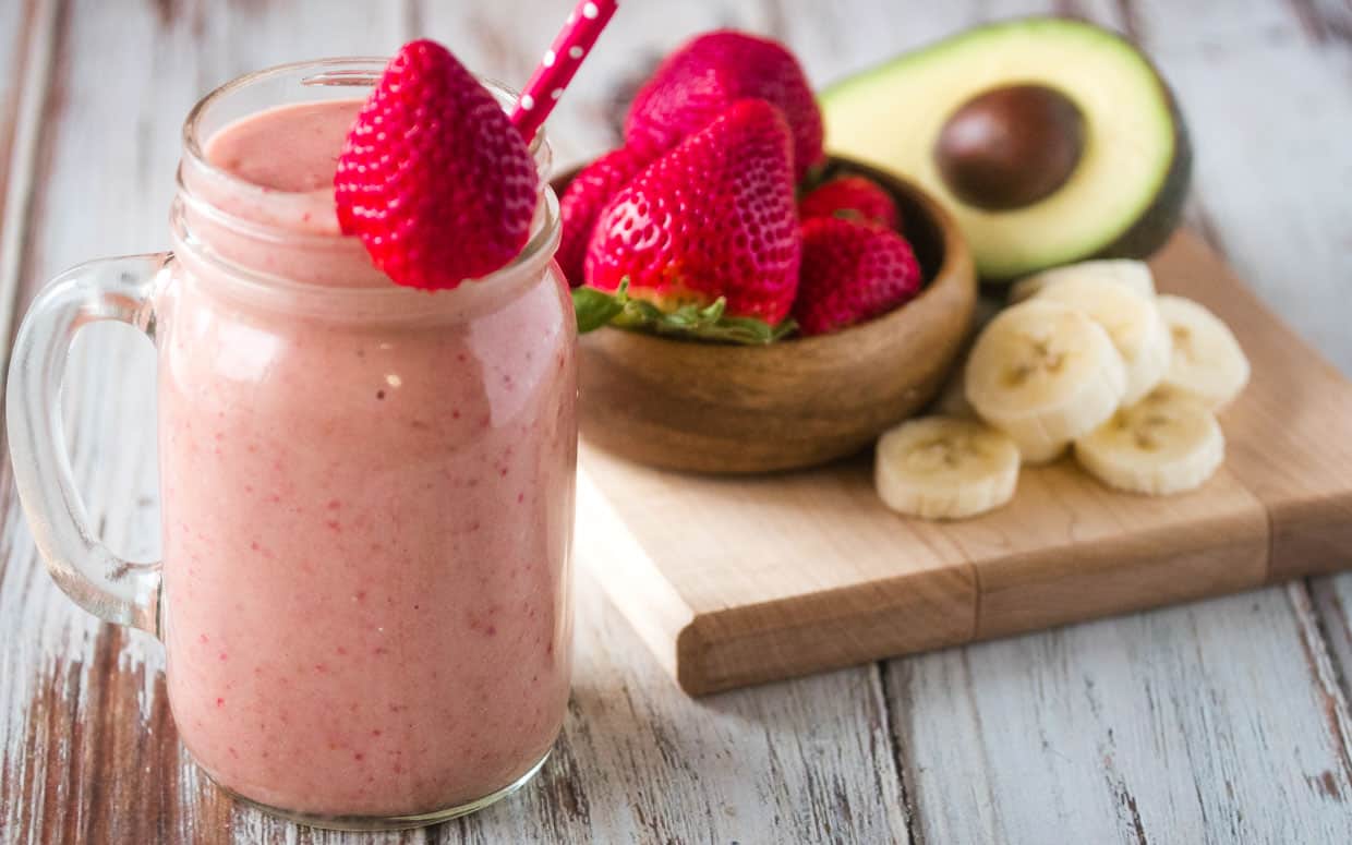 Mug of strawberry smoothie with avocado and banana topped with a strawberry and a red straw.