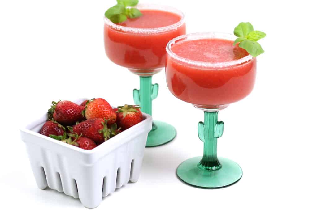 Two strawberry basil margaritas in cactus glasses with a pint of fresh berries on a white background.