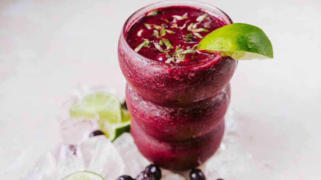 A tall clear glass filled with a frosty purple beverage garnished with fresh lime.