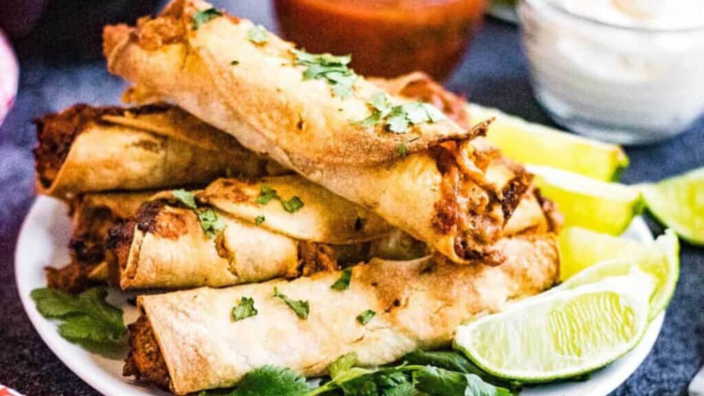Pile of taquitos on a plate with lime wedges.