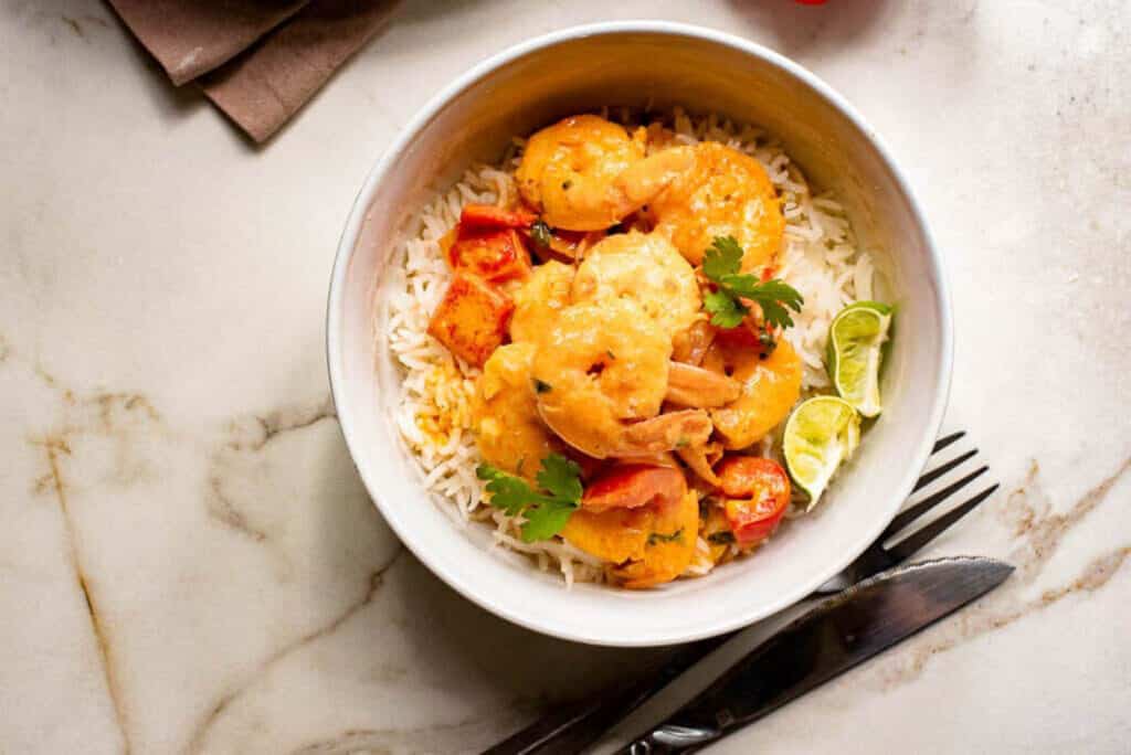 Overhead shot of a bowl of Thai shrimp curry over rice with silverware and lime wedges on the side.