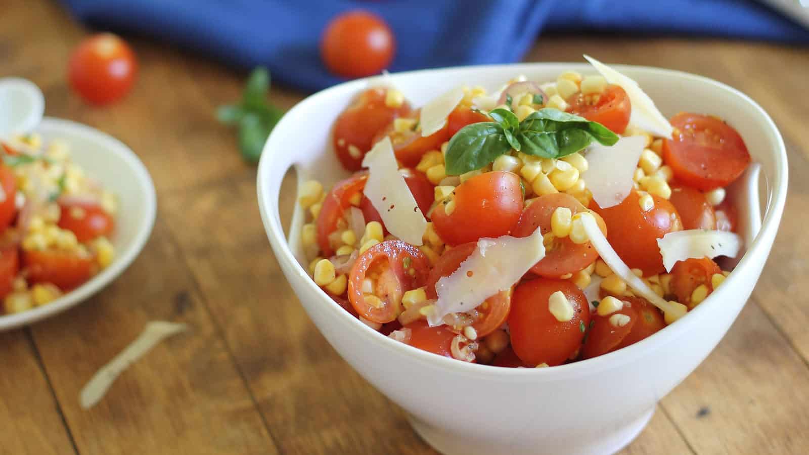 Tomato corn salad with shaved parmesan and basil garnish in white bowl.