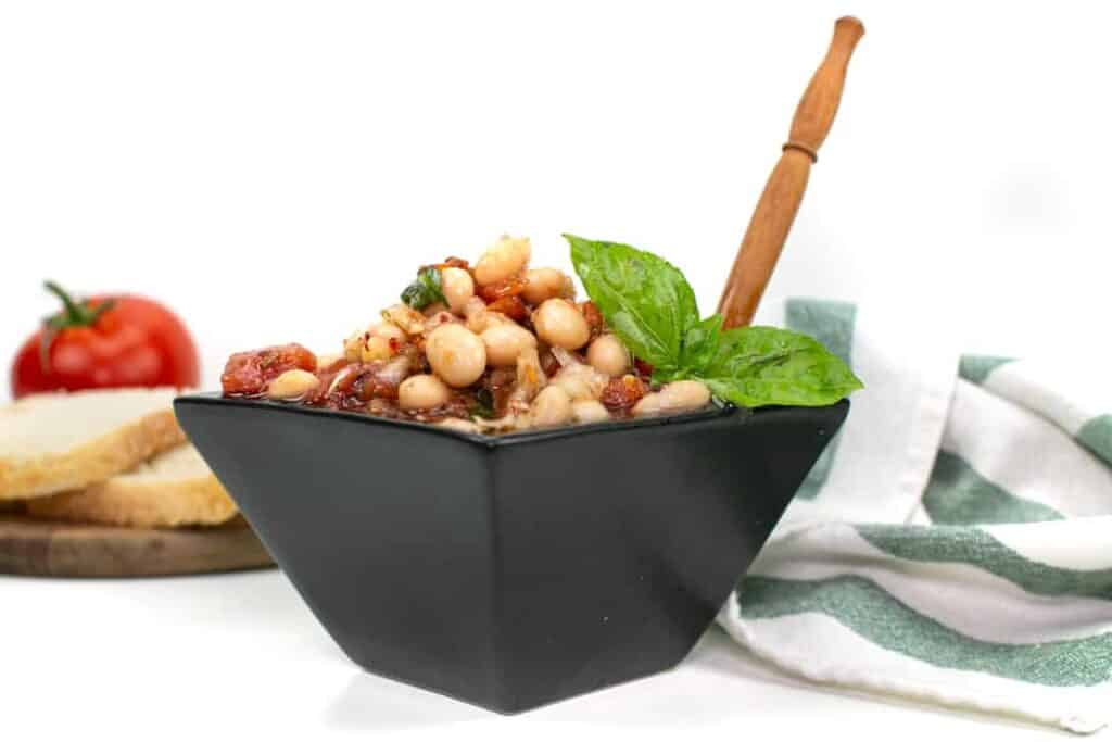 A teal towel, plate of bread and a tomato lie next to a black bowl of tuscan white bean dip.