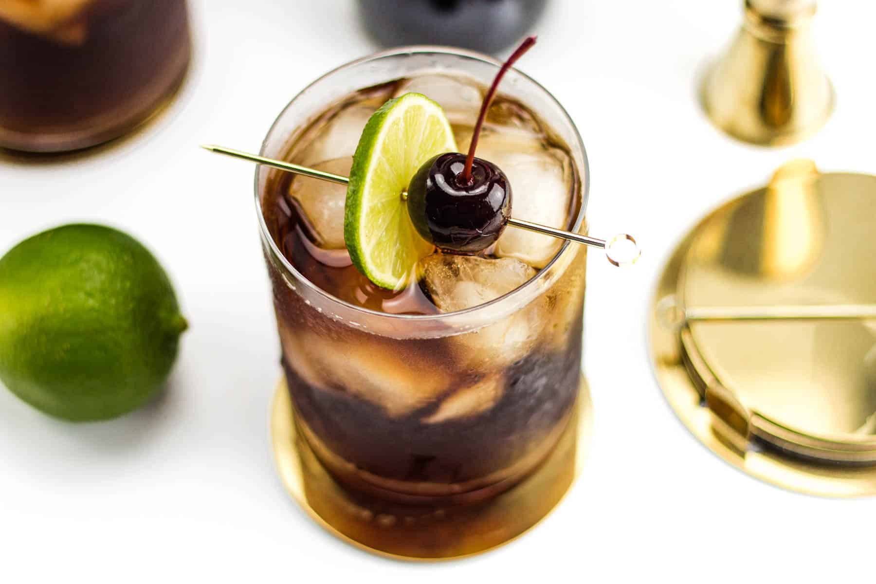 A dark brown cocktail with ice sits surrounded by a whole lime and gold cocktail tools.