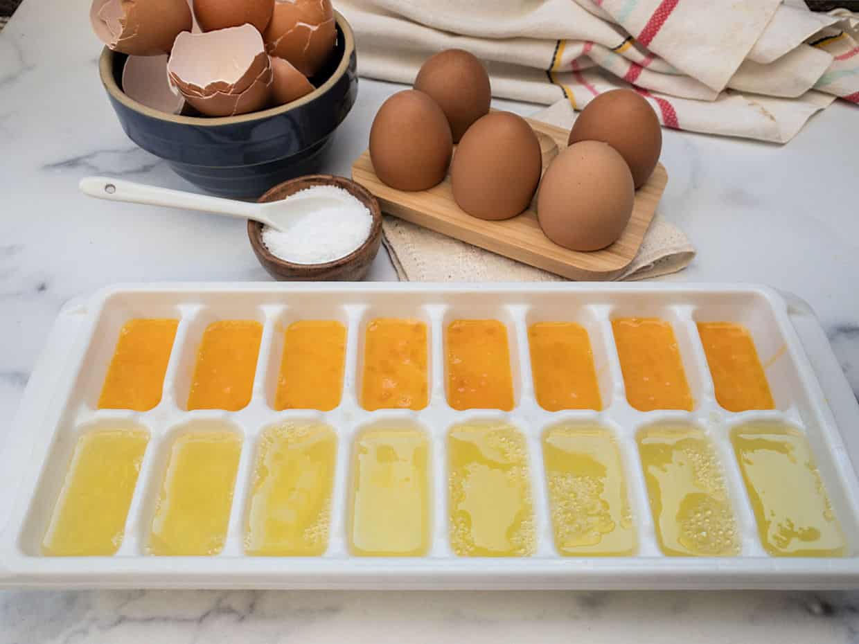 A plastic ice cube tray filled with egg whites and egg yolks with broken eggs behind it.