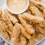 Vegan breaded zucchini fries with dipping sauce on a white plate.