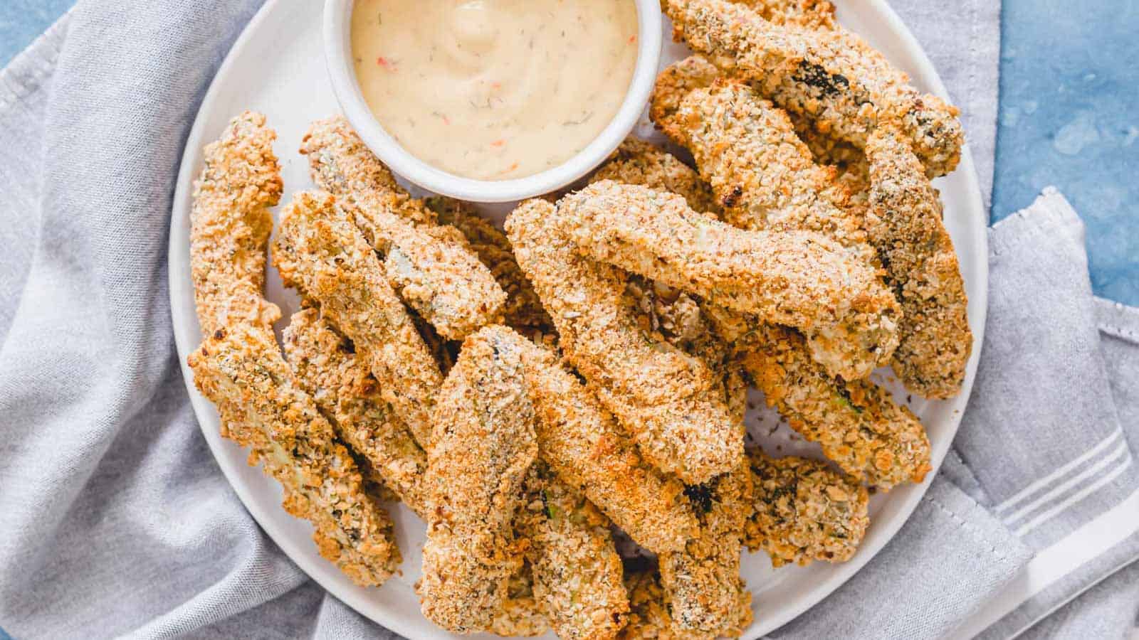 Vegan breaded zucchini fries with dipping sauce on a white plate.