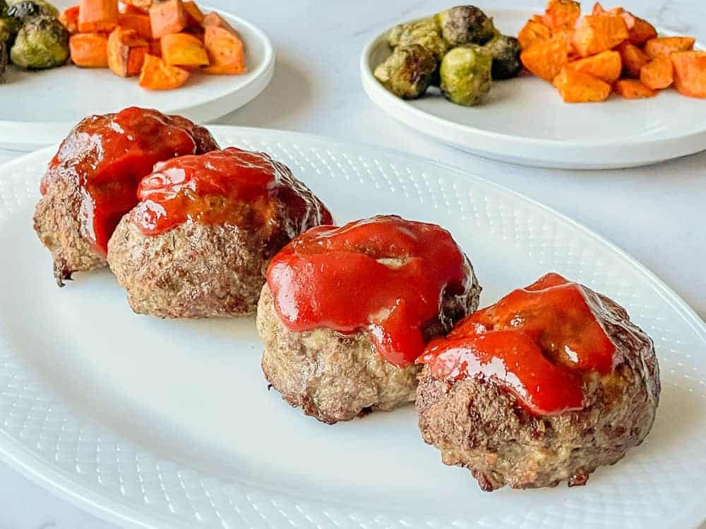 Mini Meatloaves on a white platter with sweet potatoes and brussels sprouts behind it.