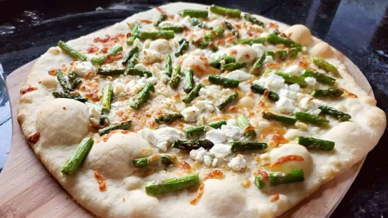 Image shows a close up of an Asparagus and Goat Cheese Pizza on a pizza peel sitting on a black counter.