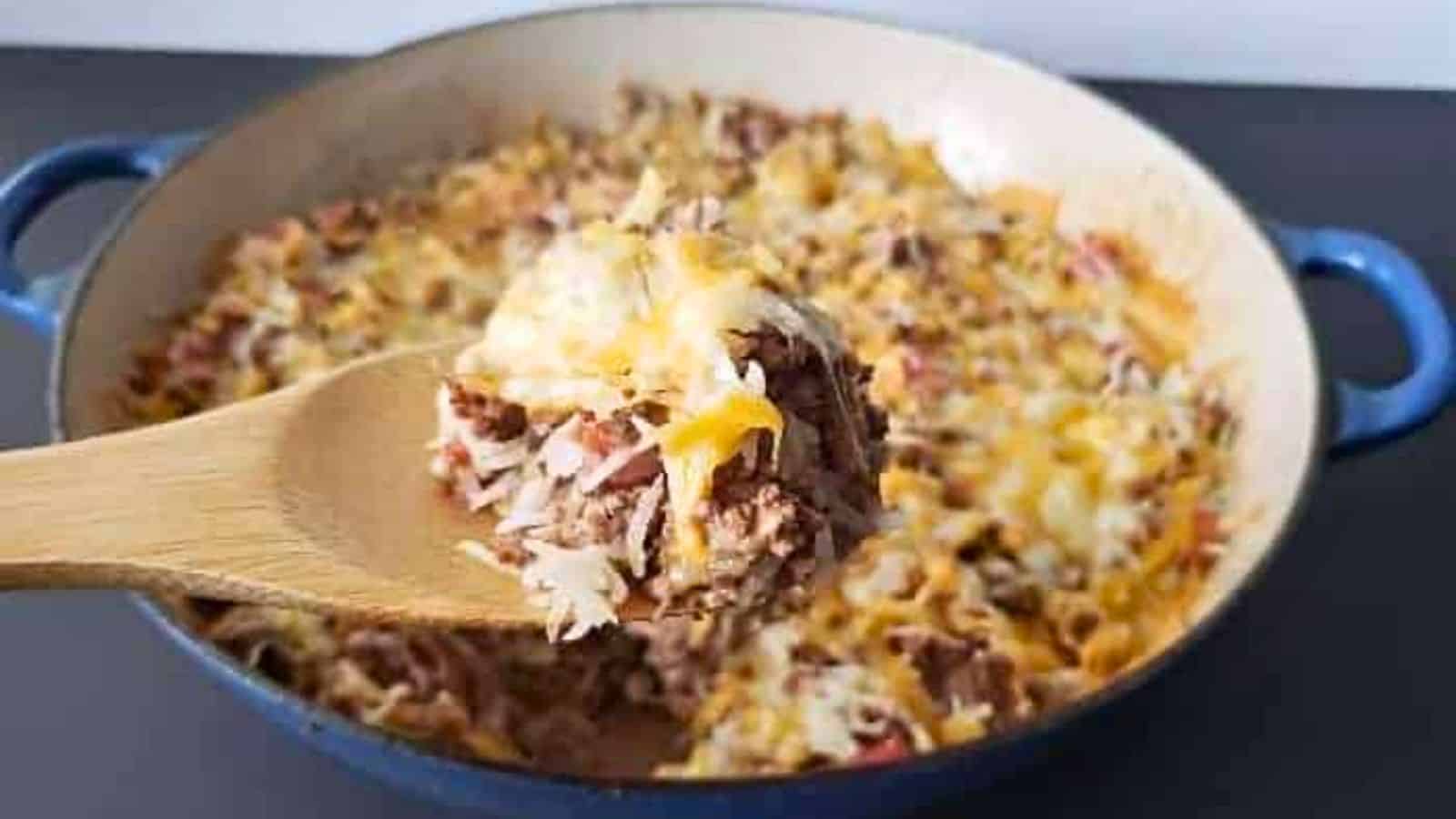 Image shows a closeup of a spoon holding some Cheesy Beef and Rice Casserole with the full skillet behind it.