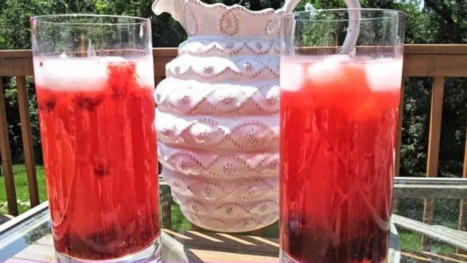 Image shows two glasses of Berry Lemonade with ice and berries at the bottom with a white pitcher behind it.