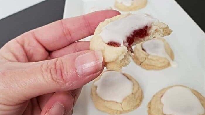 Hand holding a thumbprint cookie with a bite taken out of it over a tray filled with more classic thumbprint cookies.