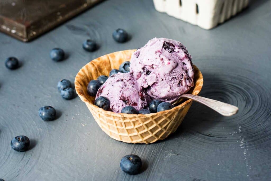 A serving of blueberry ice cream in a waffle cone bowl on a gray background. 