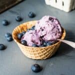 A serving of blueberry ice cream in a waffle cone bowl on a gray background.
