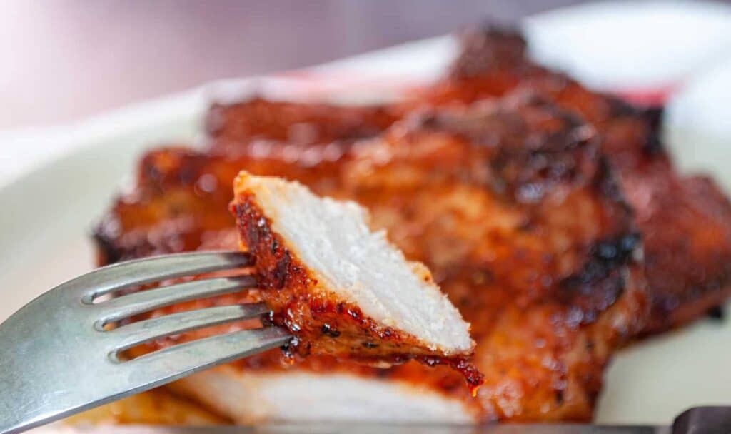 A slice of a bbq pork chop on a fork the rest of the chop is in the background.