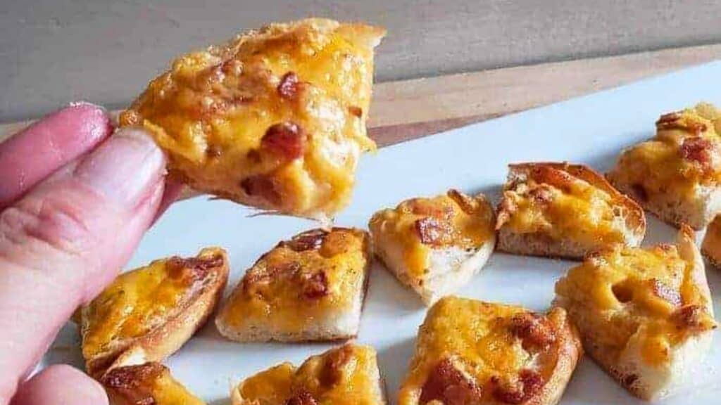 Image shows a hand holding a quarter of a Boozy Bacon Cheddar Bite with more on a white tray in the background.