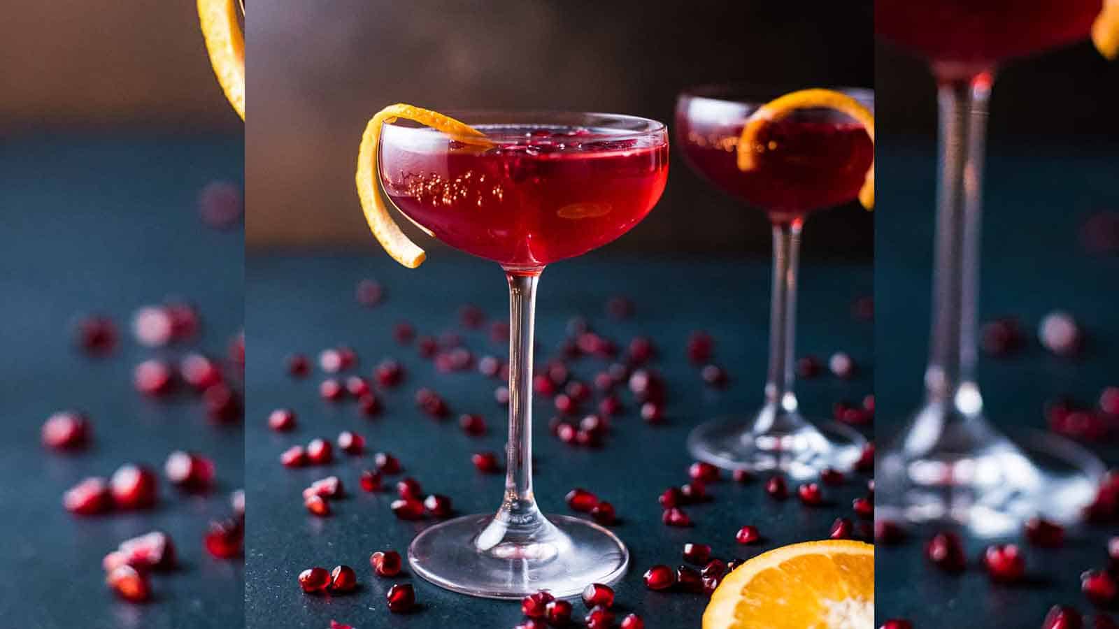 Two champagne pomegranate cocktails in coupe glasses on a dark blue background scattered with pomegranate seeds.