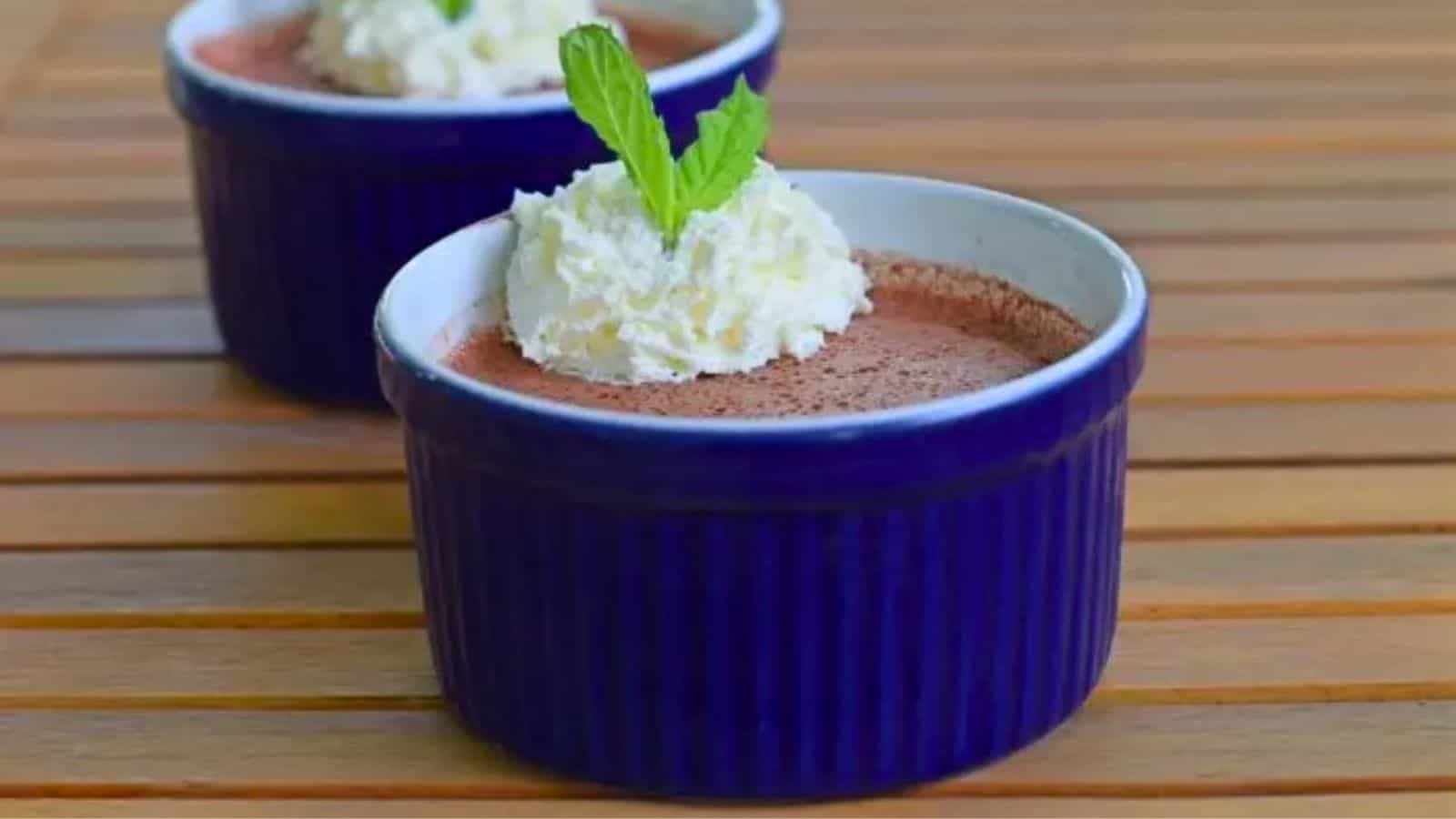 Image shows two Cherry Bavarian Custards in blue ramekins on a wooden table with whipped cream and a mint sprig.