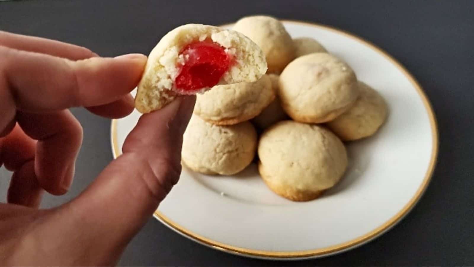 Image shows a hand holding a Cherry Surprise Cookie with more on a white plate behind it.