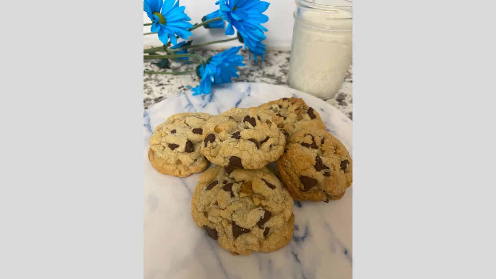Pile of chocolate chip cookies on marble plate with glass of milk in background.