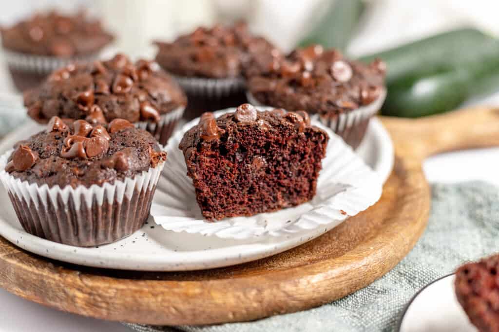 Chocolate muffin cut in half on a white plate surrounded by more muffins and zucchini.