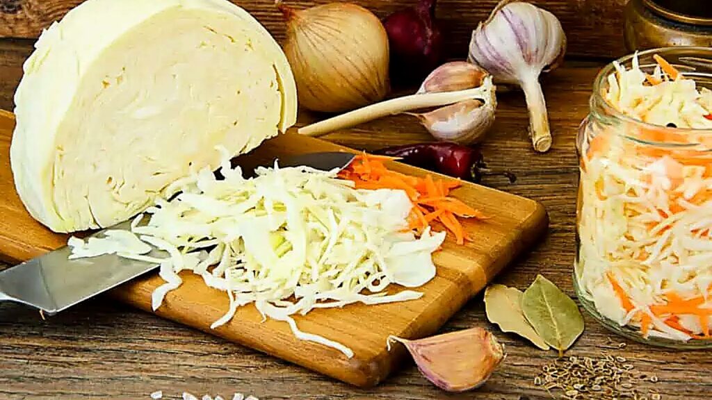 Chopped cabbage and carrots on a cutting board with a knife, surrounded by onions, garlic, spices, fermenting supplies, and a jar of shredded vegetable mix on a wooden table.