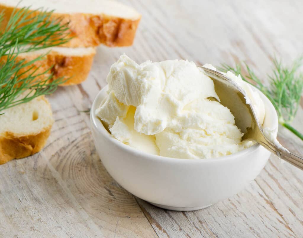 A white bowl filled with creamy white cheese&mdash;perfect for learning how to soften cream cheese&mdash;sits with a spoon, next to slices of bread and sprigs of dill on a wooden surface.
