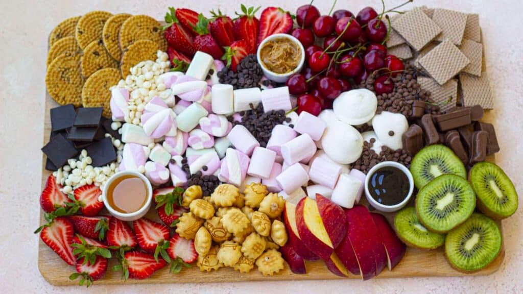 A visually captivating S'more Charcuterie Board, featuring an array of pink and white marshmallows, rich chocolates, fruits and crunchy graham crackers arranged artfully on a wooden board.