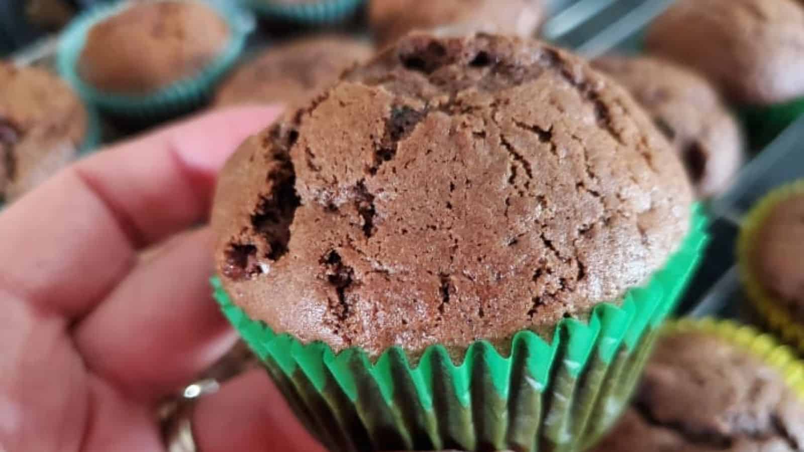 Image shows a hand holding a Double Chocolate Zucchini Muffin with more in the background.