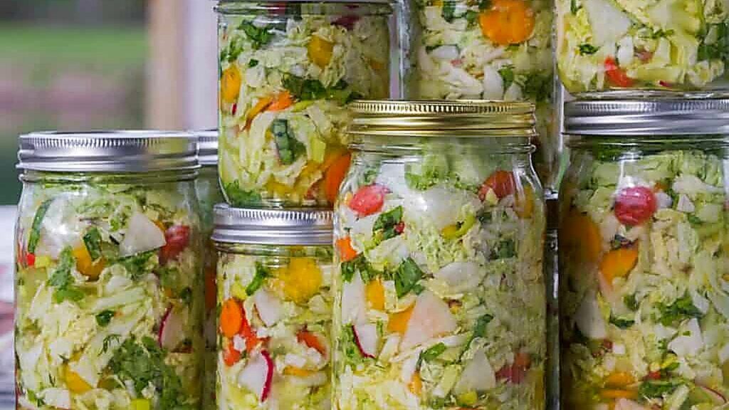 Several glass jars filled with homemade mixed vegetable pickles, including cabbage, carrots, peppers, and leafy greens, are stacked on a table alongside essential fermenting supplies.