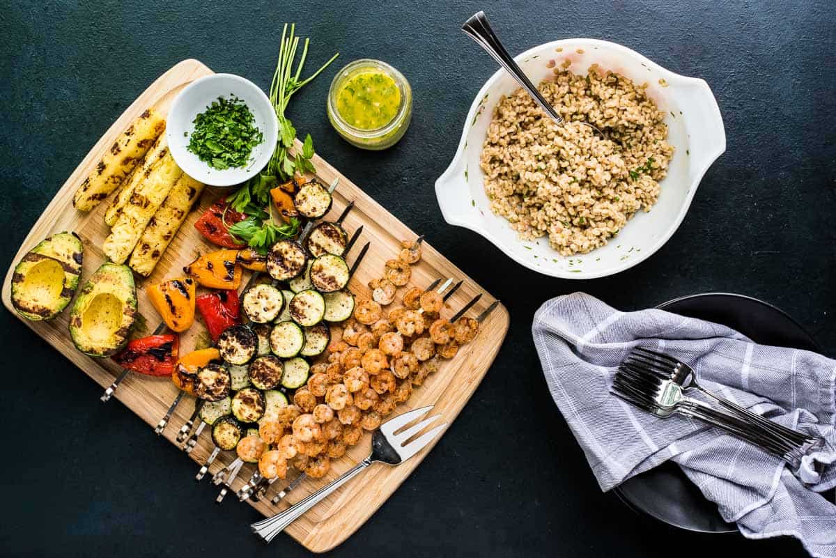 Overhead shot of shrimp skewers and veggie skewers next to a bowl of dressed grains.