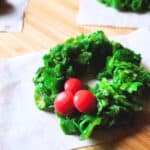 Image shows Holly Wreath Cookies on small pieces of parchment paper sitting on a wooden table.