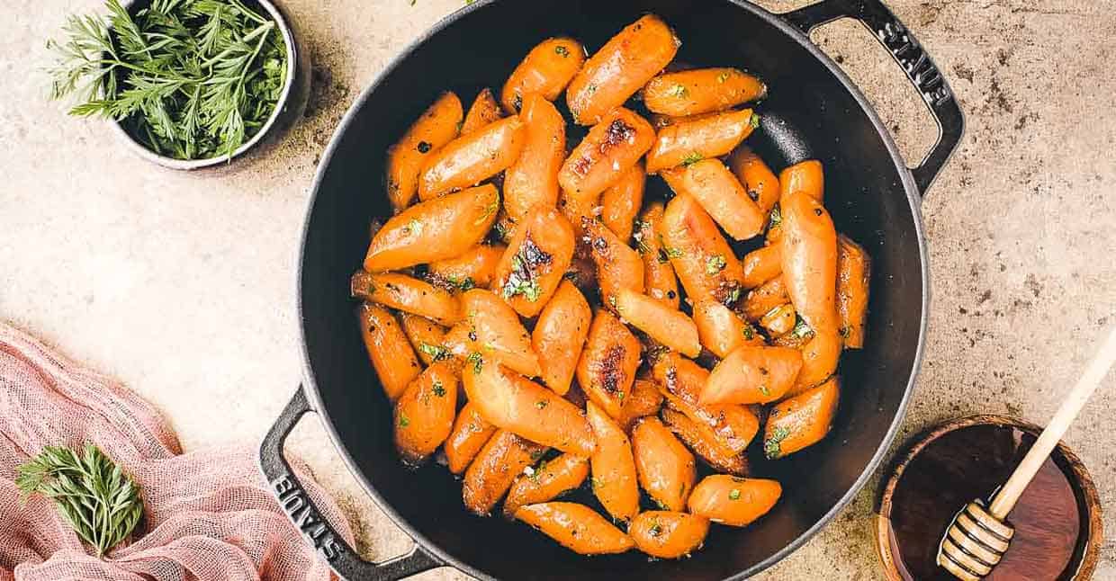 A cast iron skillet with honey garlic carrots, next to bowls of fresh herbs and honey. 