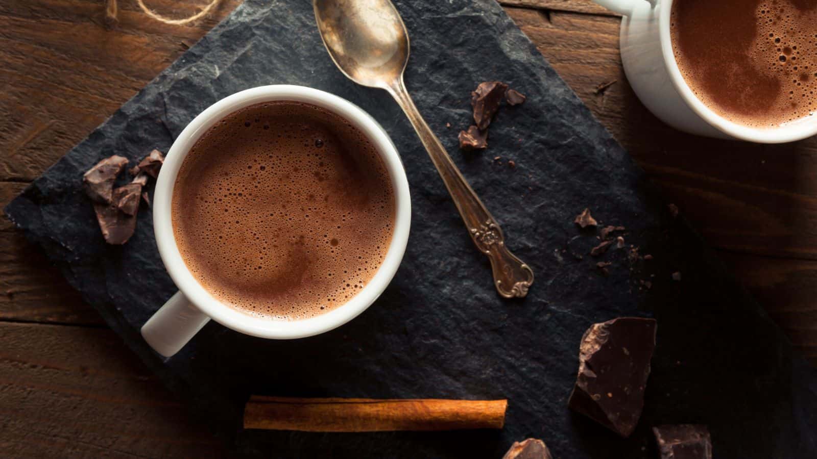 Hot chocolate in a white mug with silver spoon and ingredients around.