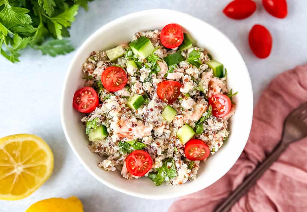 Salmon quinoa salad in a white bowl surrounded by lemons, parsley and cherry tomatoes.