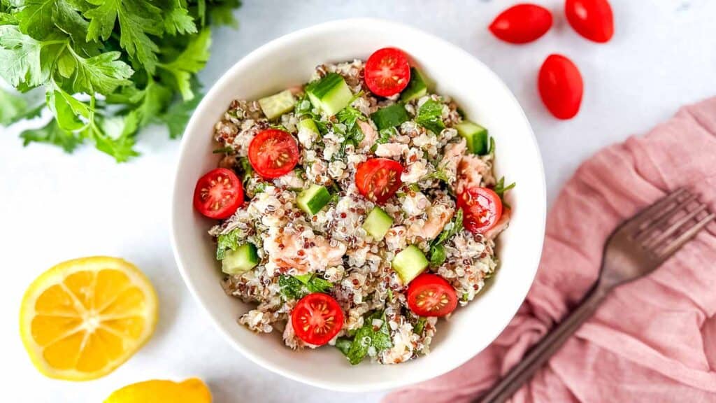 Salmon Quinoa Salad in a white bowl surrounded by parsley, cherry tomatoes, and a cut lemon.