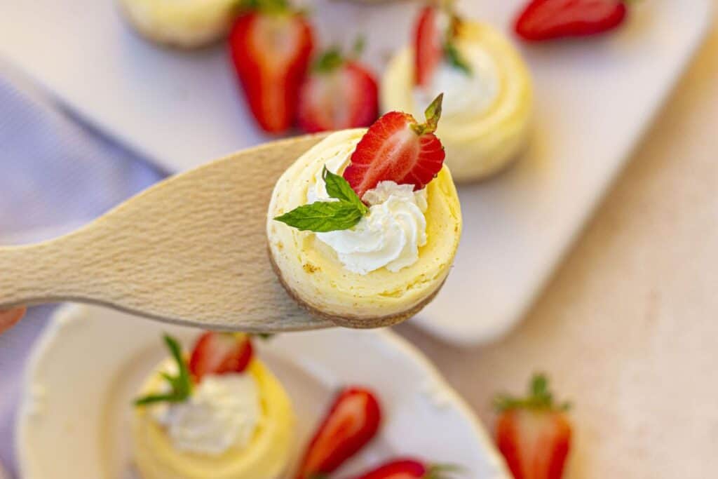 Close up of mini cheesecake on wooden spoon above plate of mini cheesecakes.