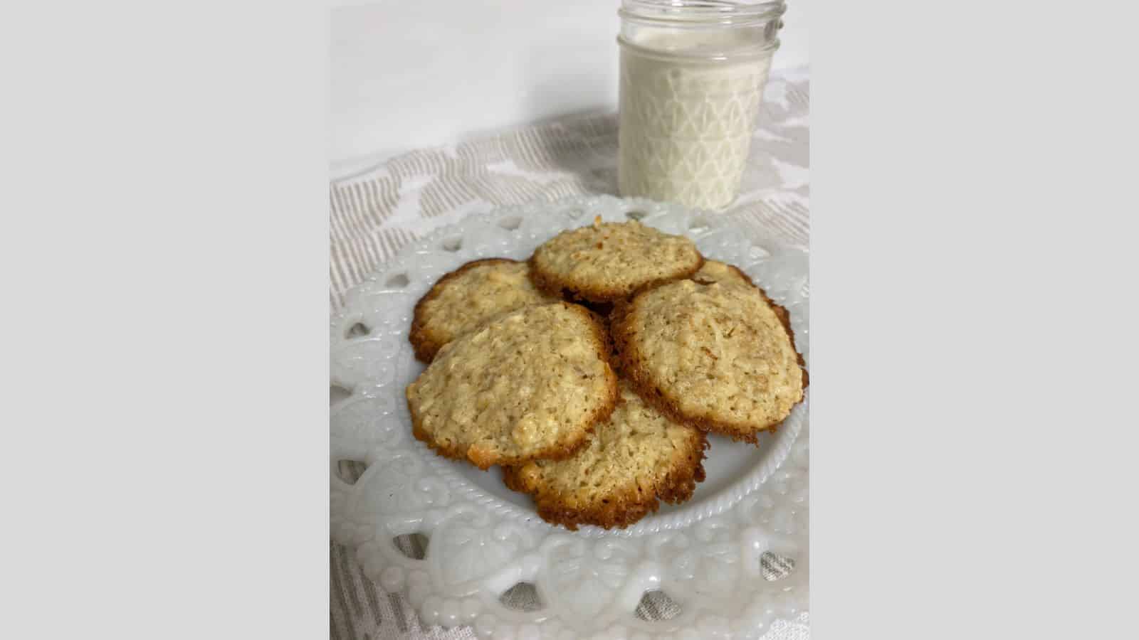 Lacy oatmeal cookies piled on white plate with glass of milk in background.