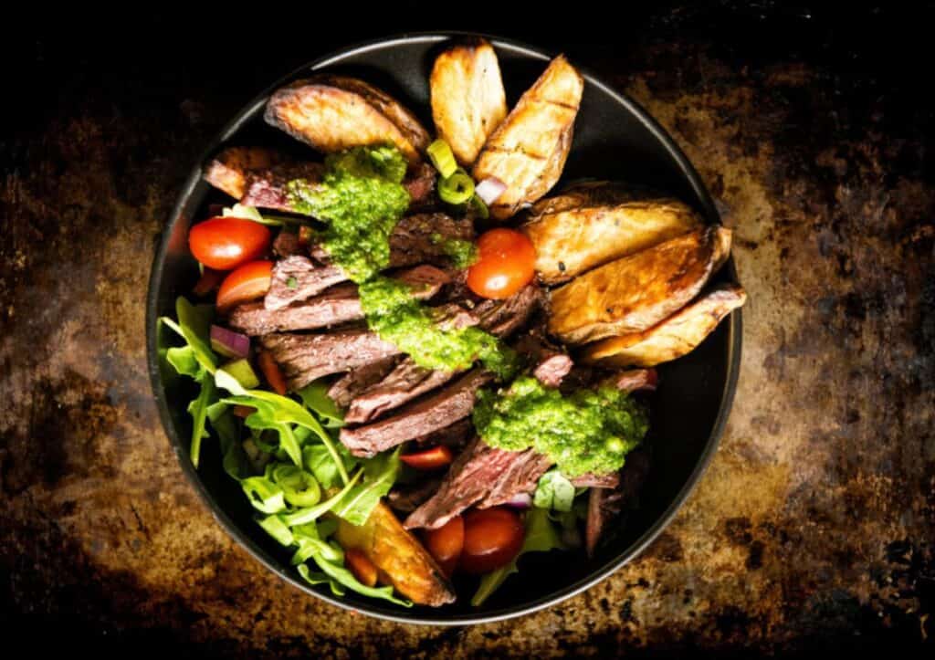 Steak, potatoes, and veggies in a bowl with chimichurri on top.