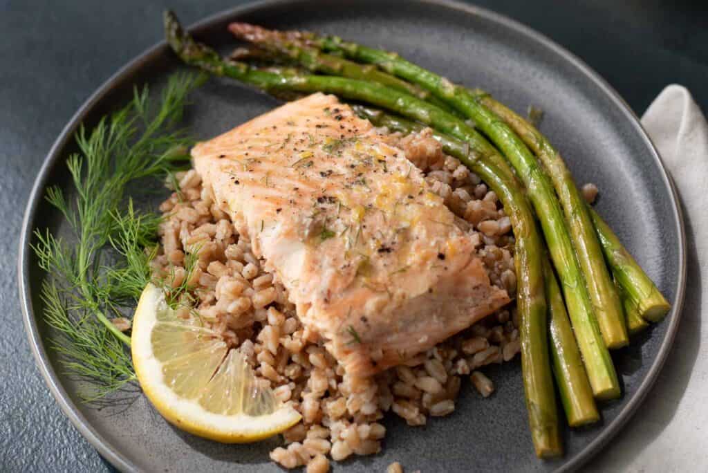 A serving of lemon dill salmon and asparagus served over faro, garnished with dill and a lemon slice.