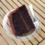 Image shows a slice of chocolate frosted chocolate cake on a plate sitting on a wooden table.