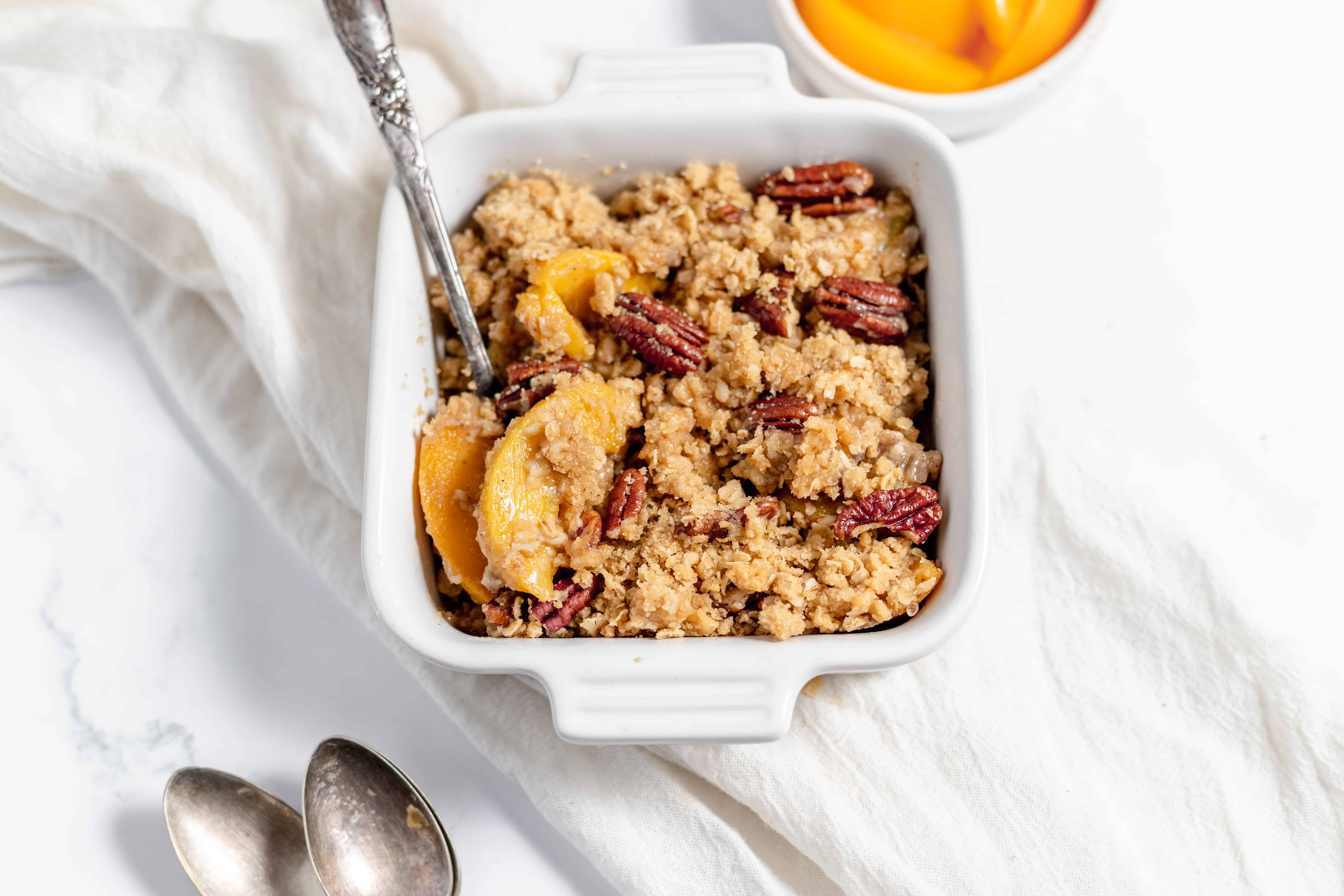 Peach crumble in a square pan with a white linen with peaches on the side. 