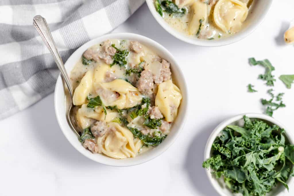 Bowls of sausage tortellini soup with spoons and a bowl of kale.