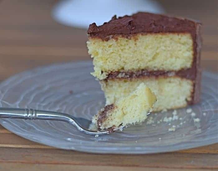 Image shows a slice of yellow cake with chocolate frosting on a plate with the full cake behind it.