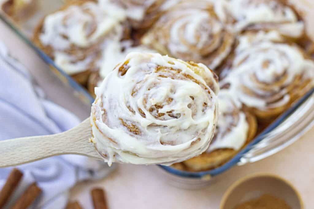 Close up of cinnamon roll on wooden spoon above dish of cinnamon rolls.